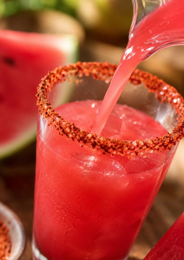 A glass filled with a bright pink watermelon drink over ice, rimmed with chili powder, is being poured into—perfect for any Agua Fresca Bar set up. A slice of watermelon and a bowl of chili powder are visible in the background.
