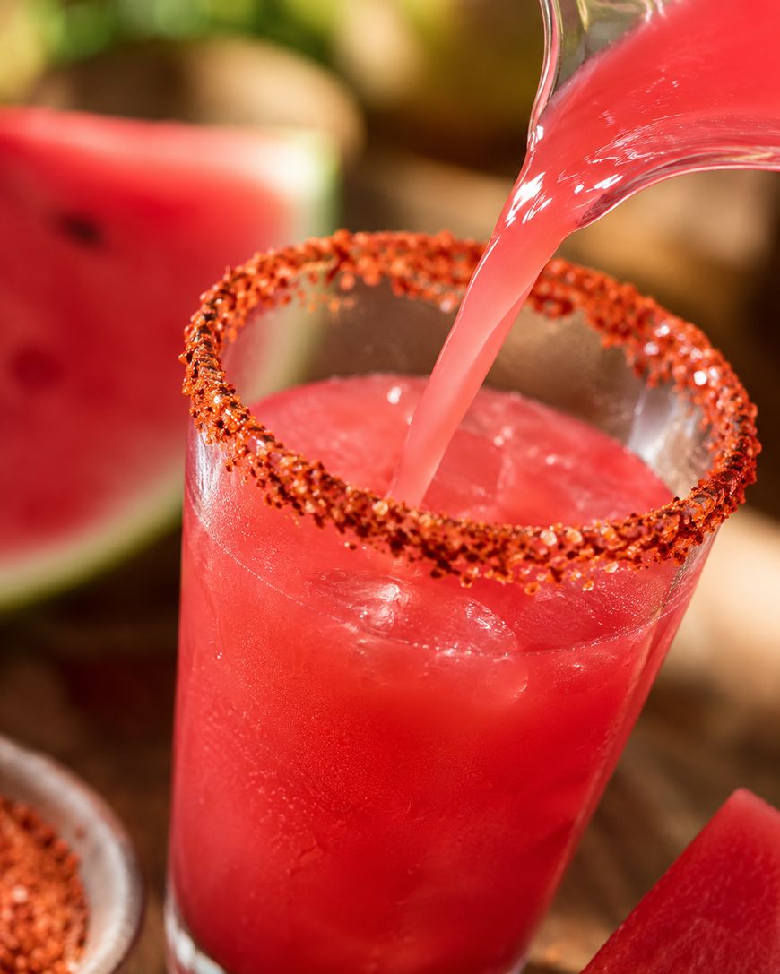A glass with a chili powder rim is being filled with a bright pink watermelon drink over ice. A slice of fresh watermelon and a bowl of chili powder are visible in the background.