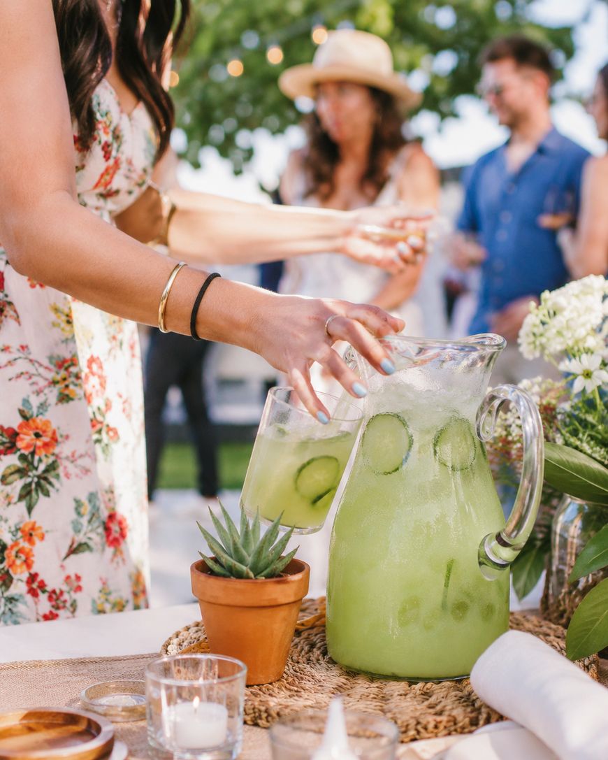 A woman in a floral dress pours a glass of cucumber-infused green drink from a large pitcher at an outdoor gathering, with people and greenery blurred in the background. A potted succulent and candles sit on the table.