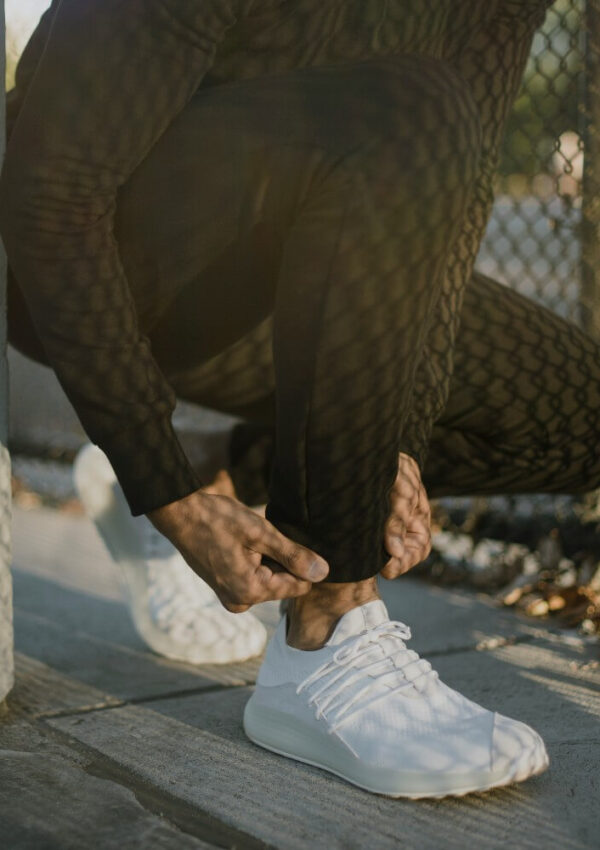 A person wearing patterned black pants and white sneakers crouches down and adjusts the cuff of their pants near a chain-link fence. Sunlight creates a shadow pattern on their clothing.