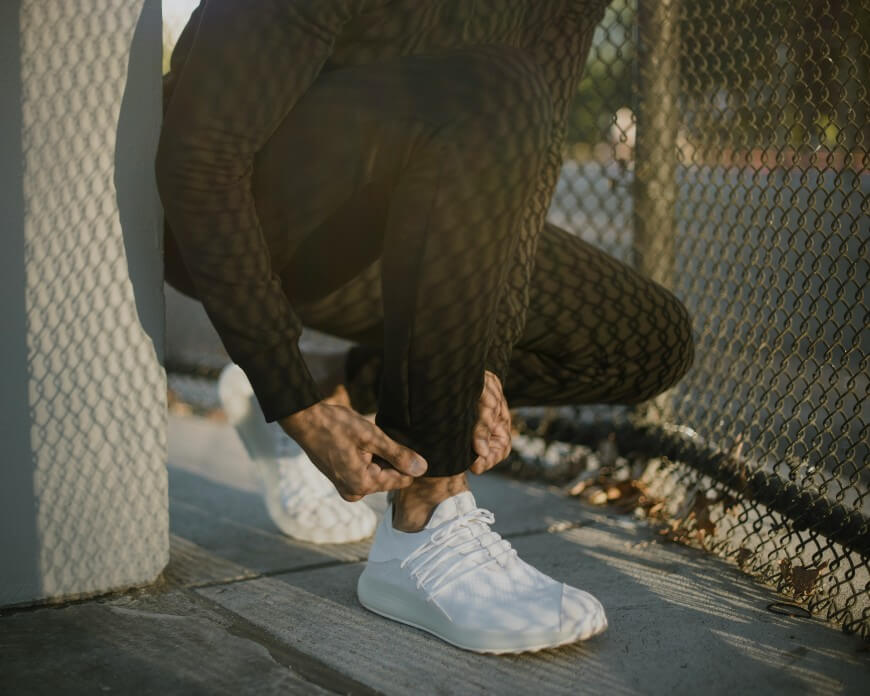 Person in patterned black athletic clothing crouches by a fence, adjusting their pant leg. They are wearing white sneakers and kneeling on a concrete surface outdoors.