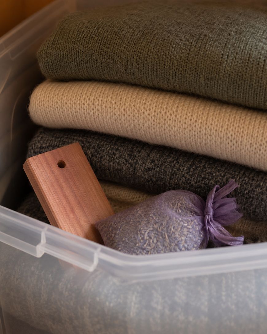 A plastic storage bin contains neatly folded sweaters in green, beige, and gray shades. Inside the bin are also a small wooden cedar block and a purple mesh sachet bag.