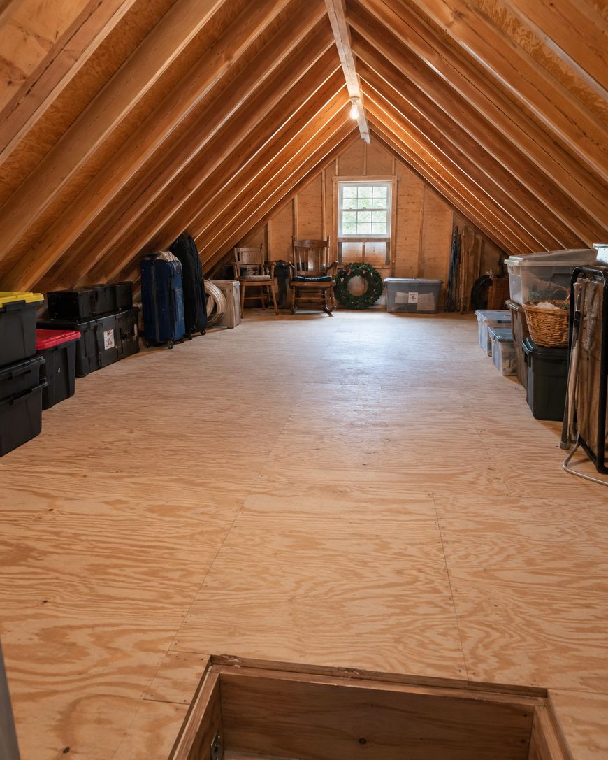 A neatly organized attic with wooden floors and exposed beams, plastic storage bins along the sides, a small window at the far end, some chairs, and a green wreath leaning against the wall.