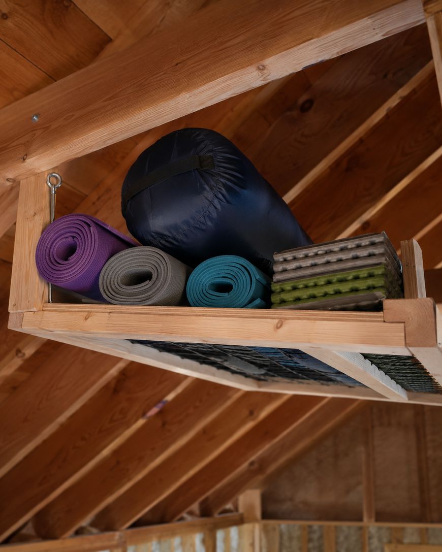 A wooden overhead shelf in a cabin stores rolled-up camping mats, a blue sleeping bag, and stackable foam pads against exposed wooden rafters and ceiling.