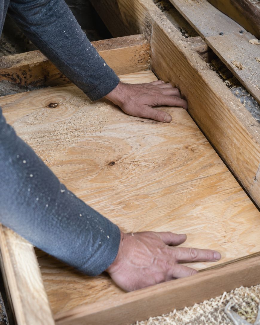 A person places a piece of plywood into a wooden frame, using both hands to adjust it; sawdust is scattered around the workspace.