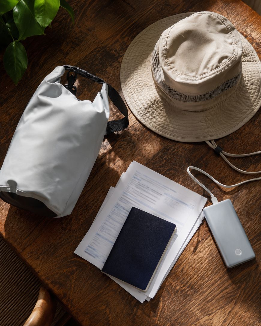 A beige sun hat, gray dry bag, black passport, papers, and a portable charger with a cable are arranged on a wooden table next to a leafy green plant in sunlight.