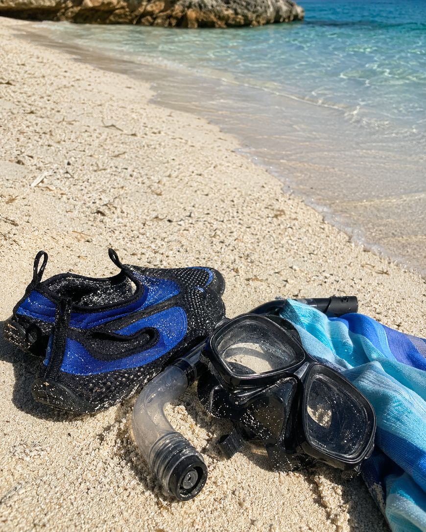 Snorkeling gear, including blue water shoes, a black snorkel mask, and a snorkel, rests on sandy beach next to a blue-striped towel, with clear ocean water and rocky shore in the background.