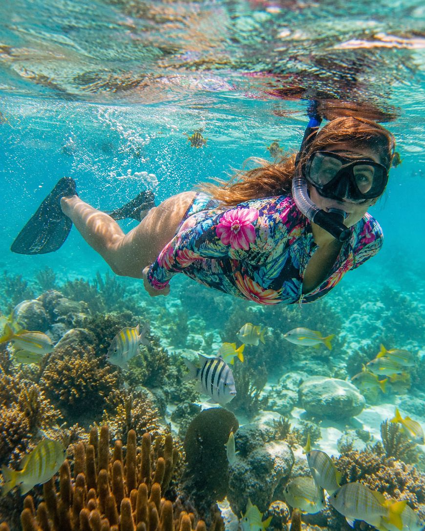 A person wearing a snorkel mask, floral swim shirt, and fins swims underwater above colorful coral reefs, surrounded by various tropical fish in clear blue water.