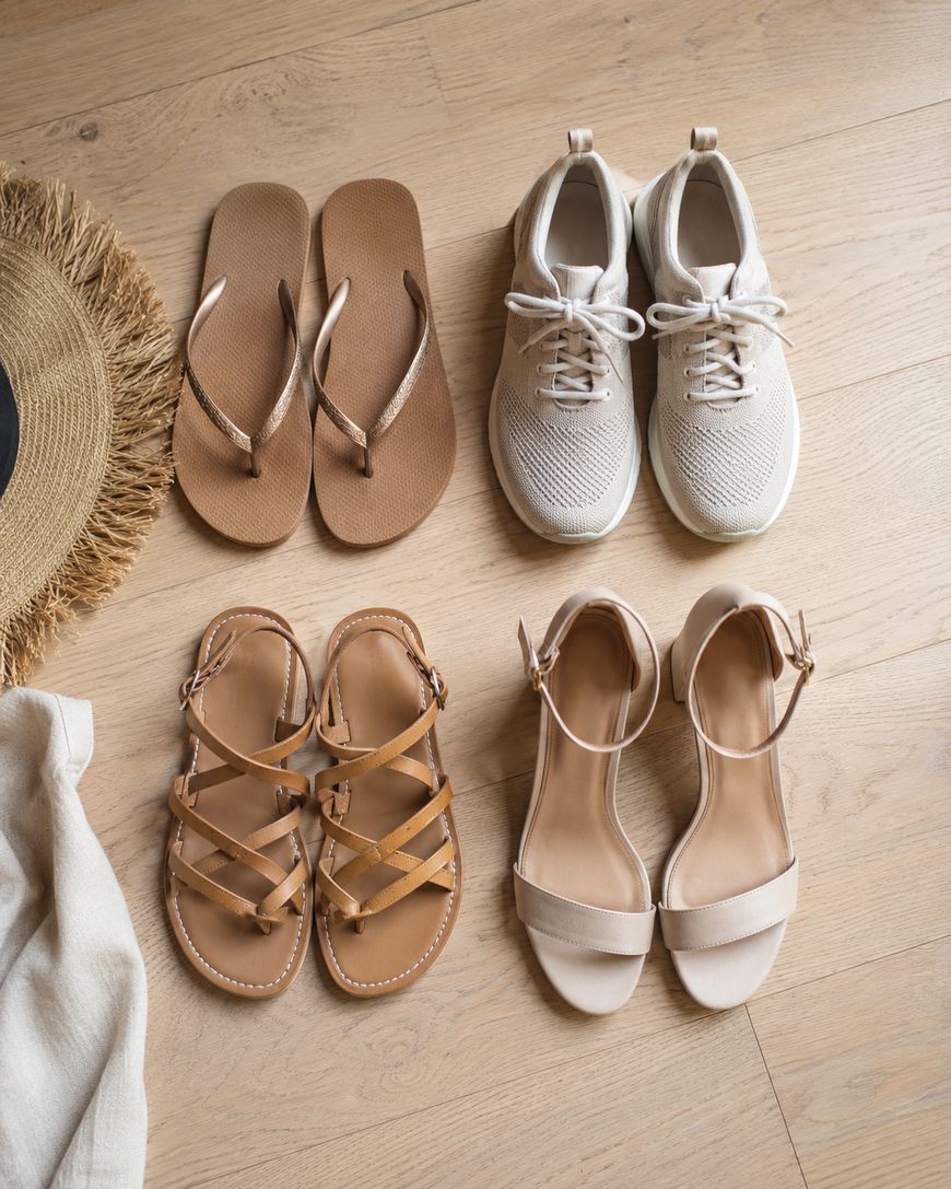 Four pairs of summer shoes—flip-flops, sneakers, strappy sandals, and heeled sandals—are neatly arranged on a light wooden floor, next to a straw hat and a light-colored cloth.