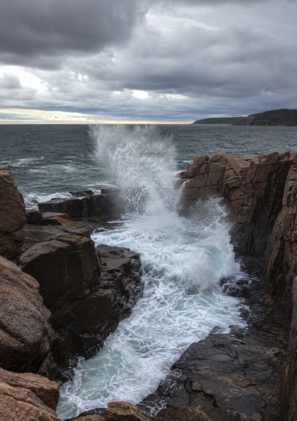 Waves crash dramatically against rugged, rocky cliffs near Bar Harbor, Maine under a cloudy sky, sending a spray of white water upward—capturing the wild beauty of Acadia visible on the distant horizon.