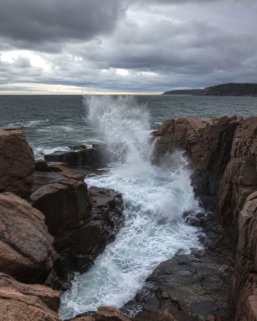 Waves crash dramatically against rugged brown cliffs under a cloudy, overcast sky, sending spray high into the air along a rocky coastline.