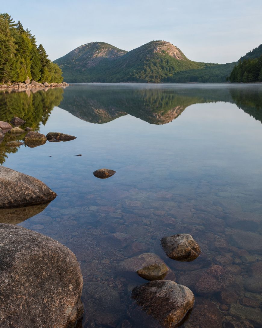 Calm lake with clear water and rocks in the foreground, reflecting two tree-covered hills and a forested shoreline under a pale blue sky.