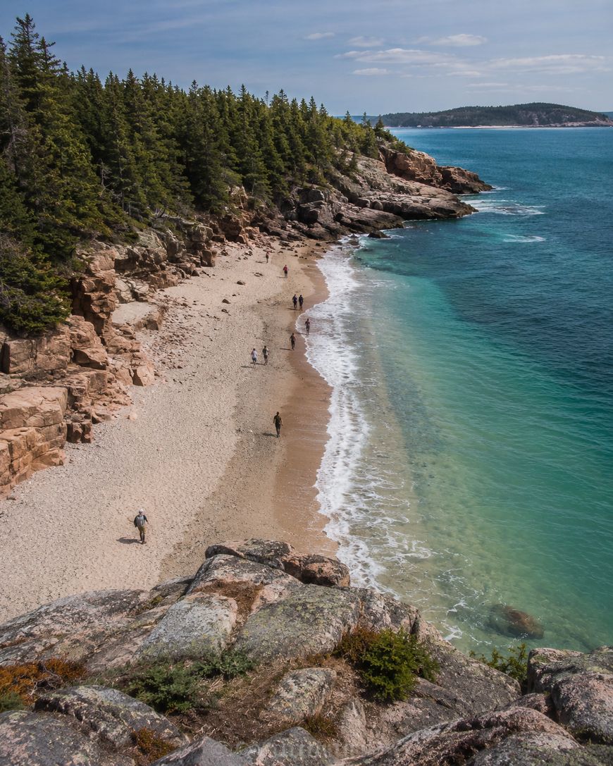 People walk along a secluded, rocky beach bordered by clear turquoise water and dense pine forest under a partly cloudy sky. Rocky cliffs rise above the shoreline, and distant land is visible across the bay.