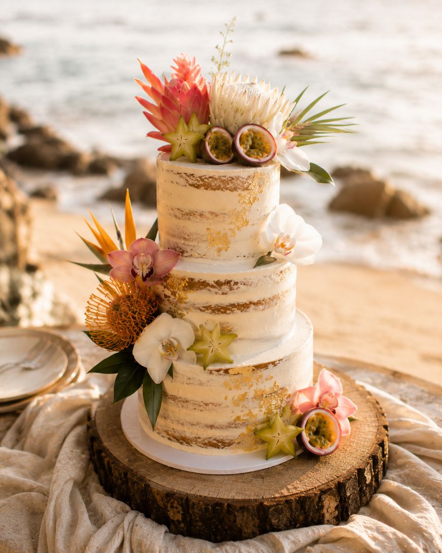A three-tiered semi-naked cake decorated with tropical flowers, sliced passion fruit, and starfruit, sits on a wooden board with a beach and ocean in the background.
