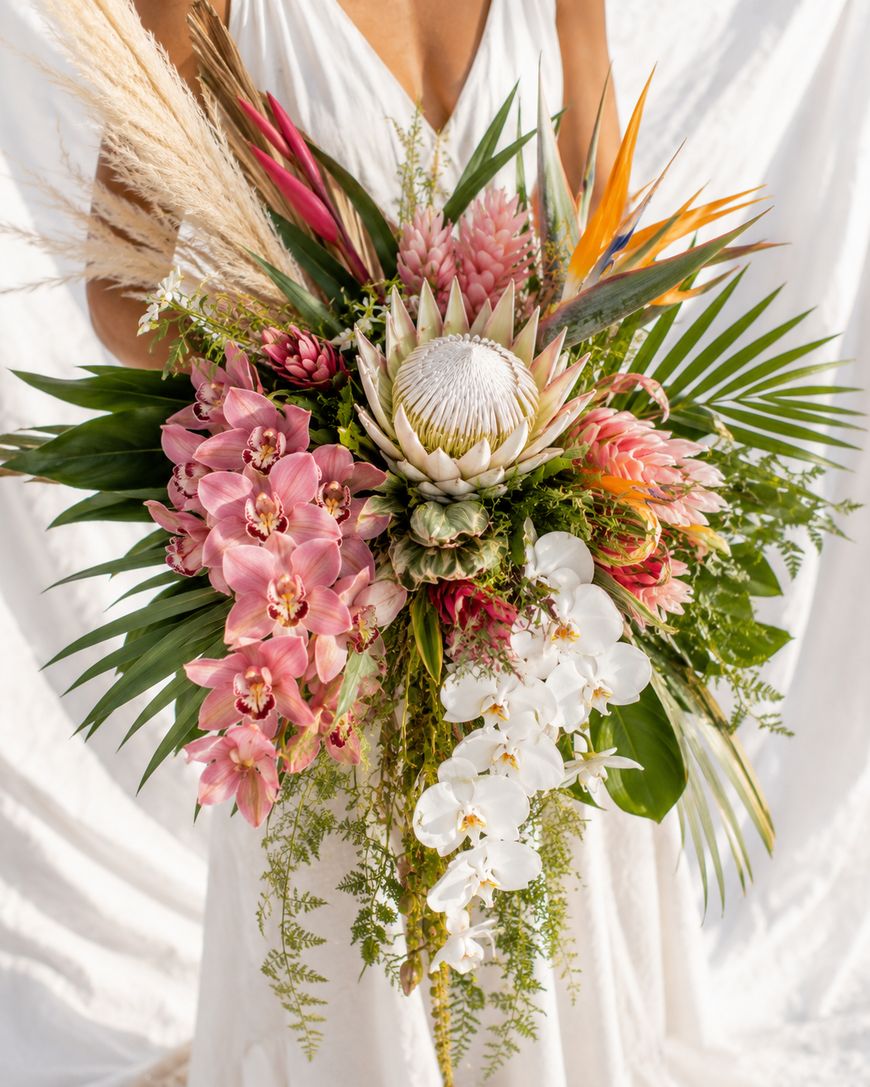 A person in a white dress holds a large, colorful bouquet featuring tropical flowers like pink orchids, white protea, ginger flowers, bird of paradise, green palm leaves, and hanging greenery.