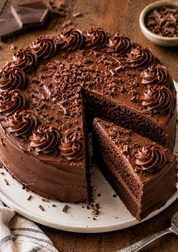 A round chocolate layer cake with rich chocolate frosting, decorated with swirls and chocolate shavings, sits on a plate with one slice cut and slightly pulled out—an irresistible treat inspired by the best Bisquick recipes. Chocolate pieces are nearby on the table.