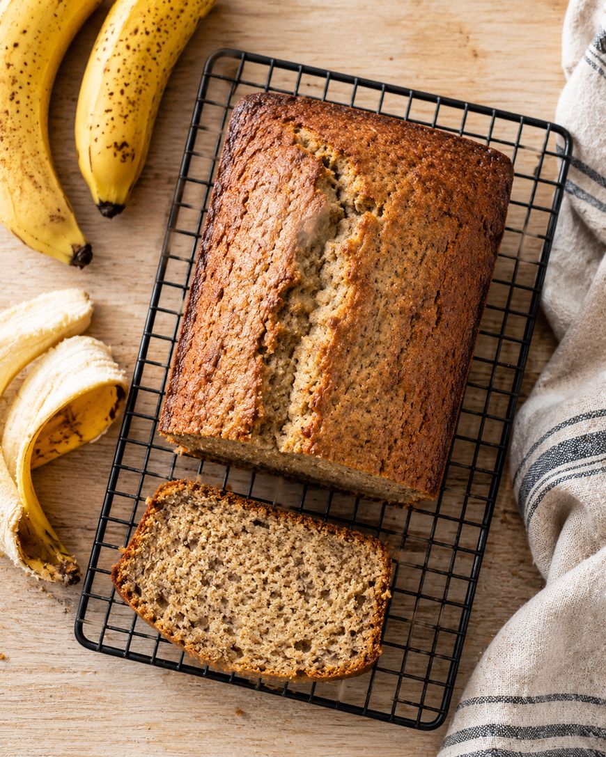A loaf of banana bread with one slice cut, placed on a cooling rack. Ripe bananas and a peeled banana lie nearby on a wooden surface with a striped kitchen towel to the side.