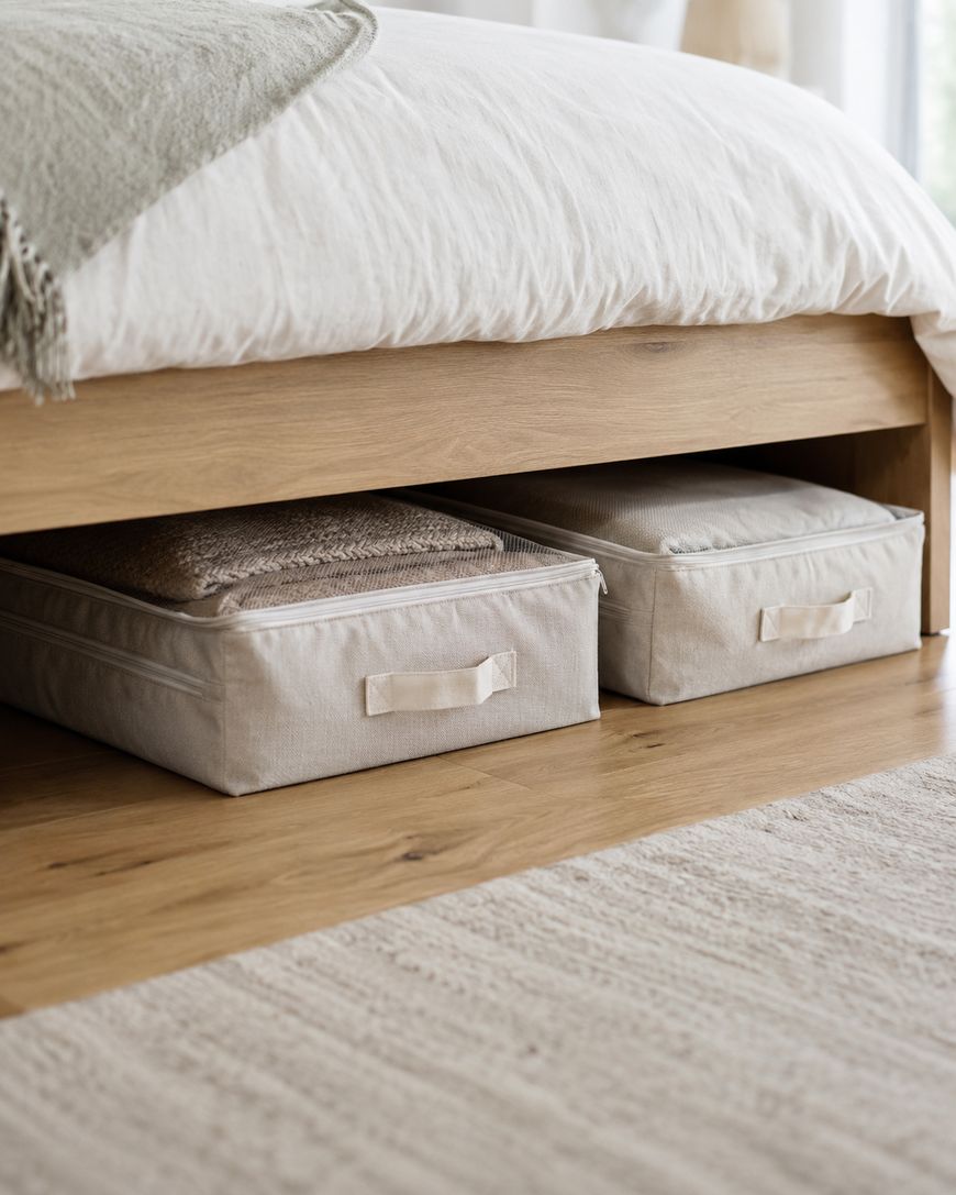 Two beige fabric storage bins with handles are neatly placed under a wooden bed, on a light wooden floor with a textured area rug nearby. The bed has white bedding and a folded blanket on top.