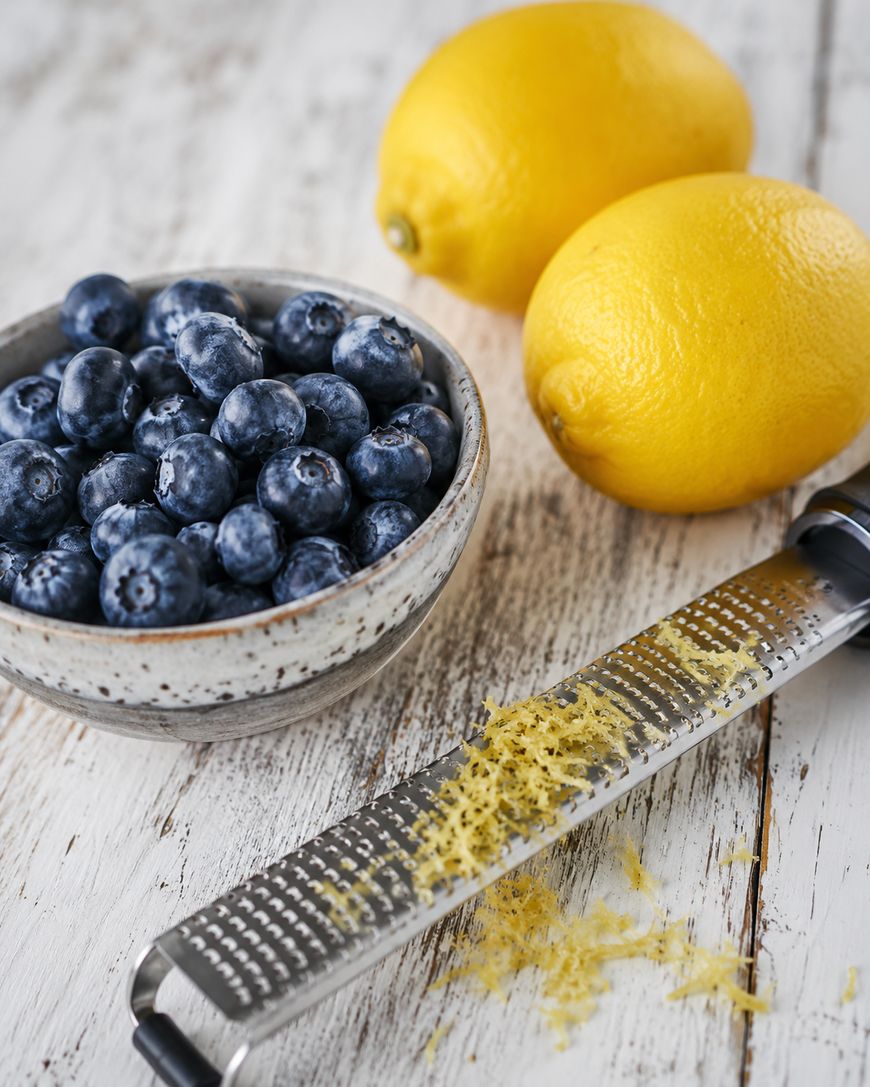 A bowl of fresh blueberries, two whole lemons, and a zester with lemon zest on a white wooden surface.