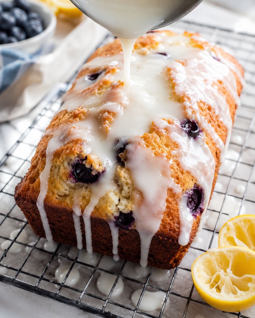A loaf of blueberry lemon bread sits on a cooling rack as thick white glaze is poured over it, with fresh lemon halves and a bowl of blueberries nearby.