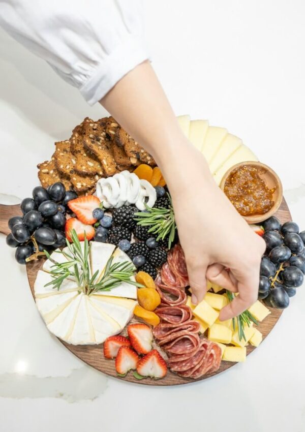 A hand reaches for cheese on a round wooden charcuterie board with salami, sliced cheeses, crackers, grapes, strawberries, blackberries, dried apricots, and a small bowl of jam, set on a white surface.