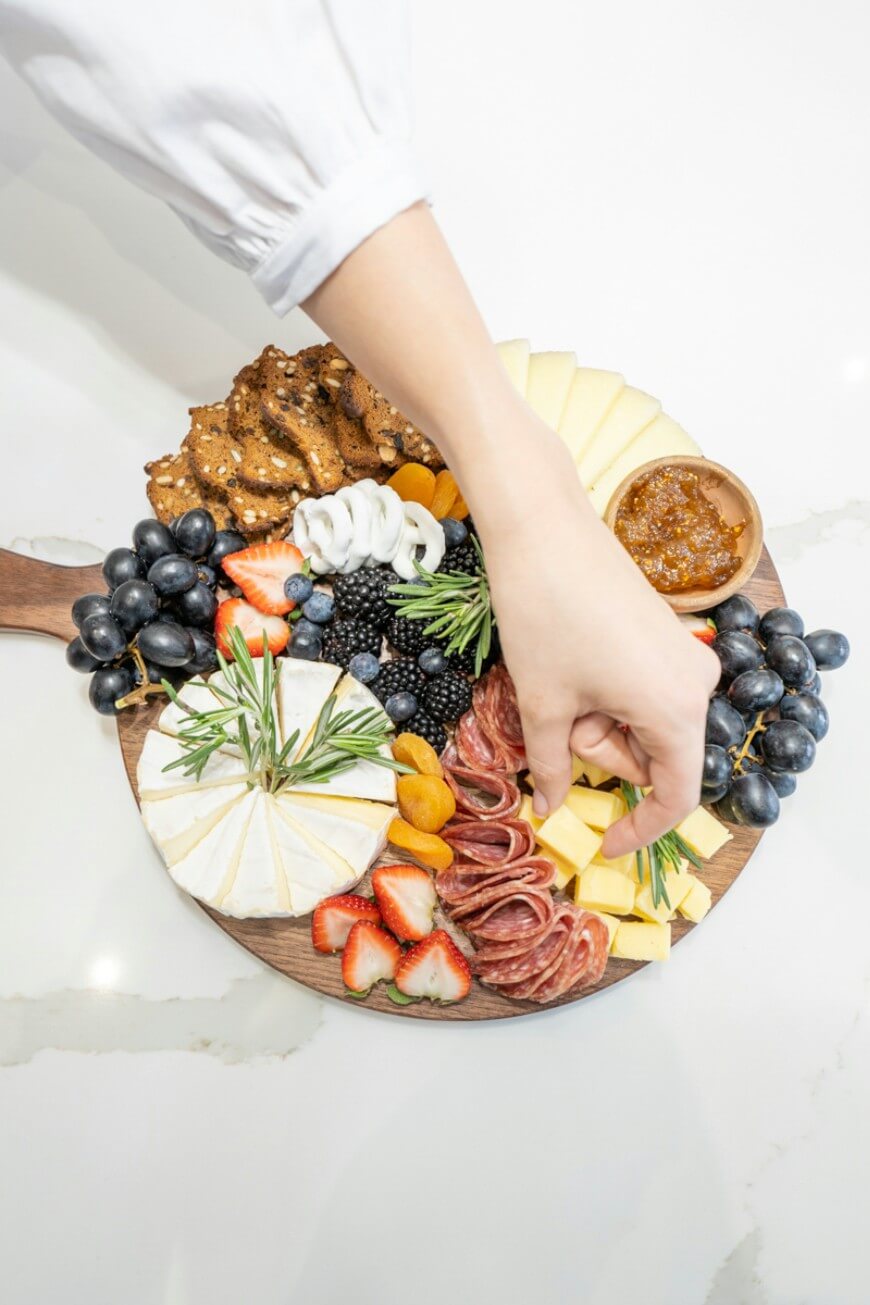 A hand reaches for cheese on a wooden charcuterie board with sliced meats, cheeses, grapes, blackberries, strawberries, crackers, dried apricots, and a small bowl of jam, arranged on a white surface.