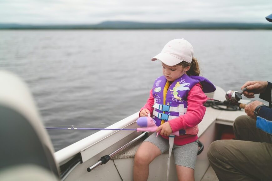 A young girl in a pink sweater, purple life jacket, and white cap sits on a boat holding a fishing rod, with an adult beside her, both fishing on a calm lake.