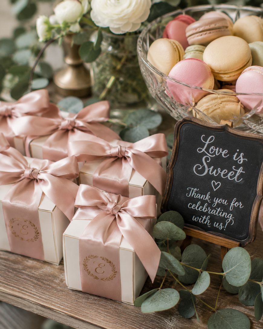 A display with pastel macarons in a glass bowl, three gift boxes with pink satin bows, white flowers, and a small chalkboard sign that reads, Love is sweet. Thank you for celebrating with us! surrounded by greenery.