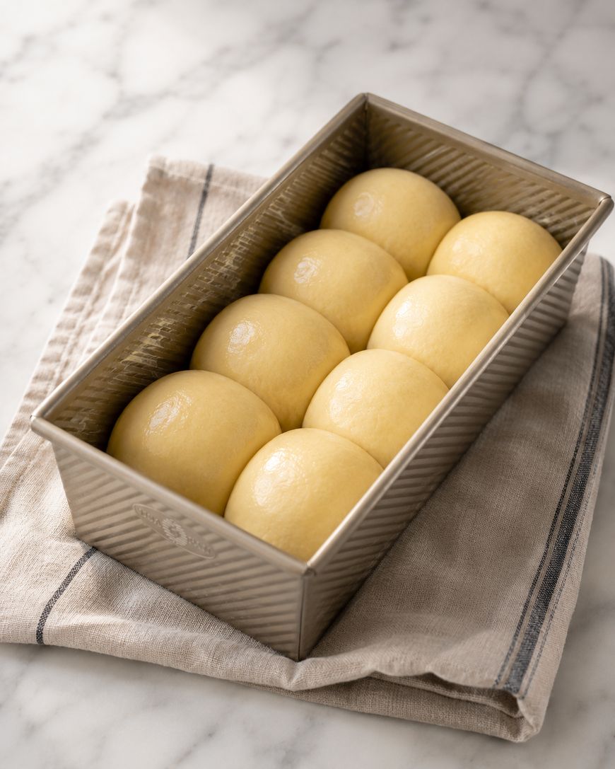 Eight smooth, round dough balls are arranged in a loaf pan, resting on a beige kitchen towel with dark stripes, set on a marble countertop.