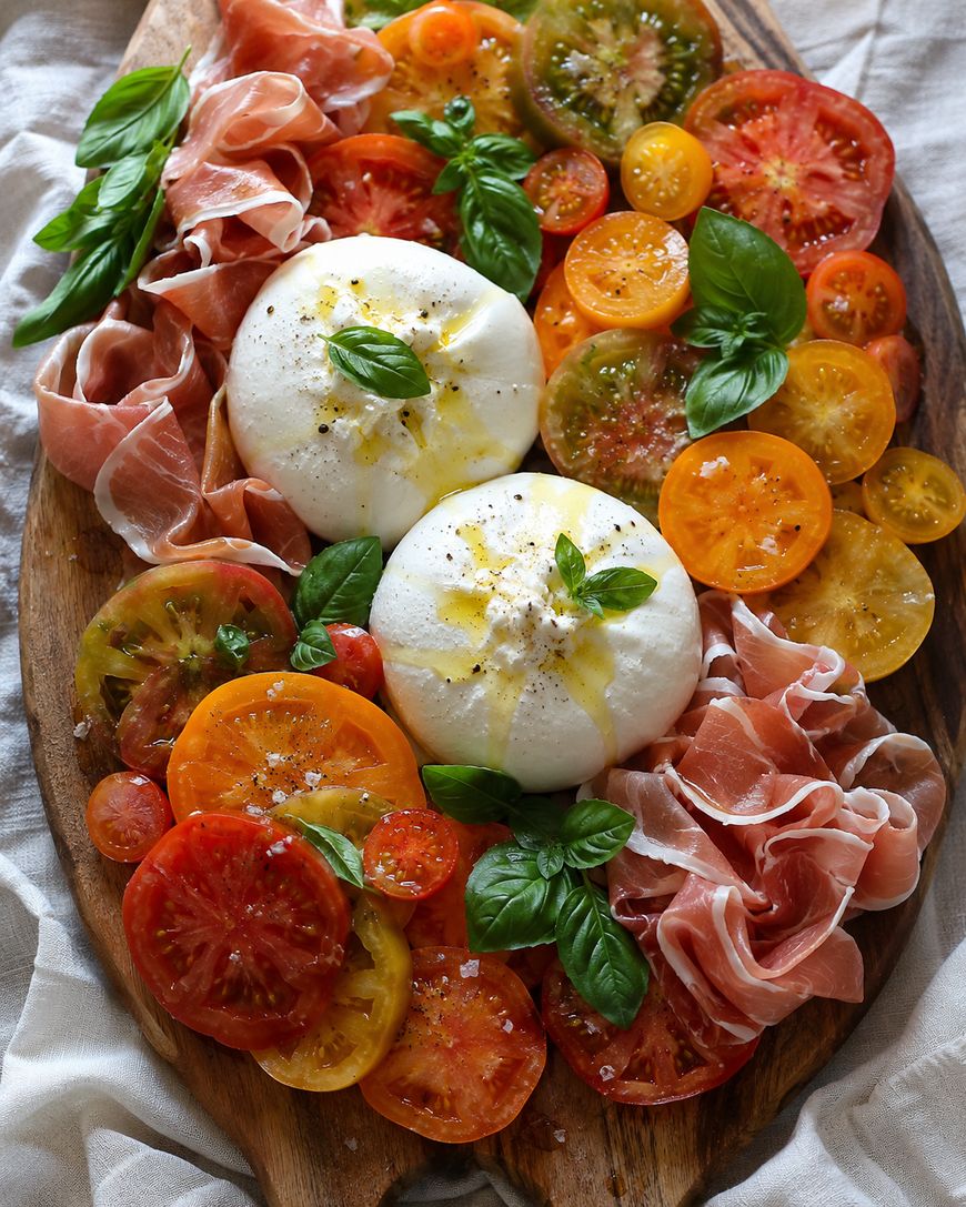 A wooden board topped with colorful heirloom tomato slices, burrata cheese drizzled with olive oil, fresh basil leaves, and thin slices of prosciutto arranged artfully.