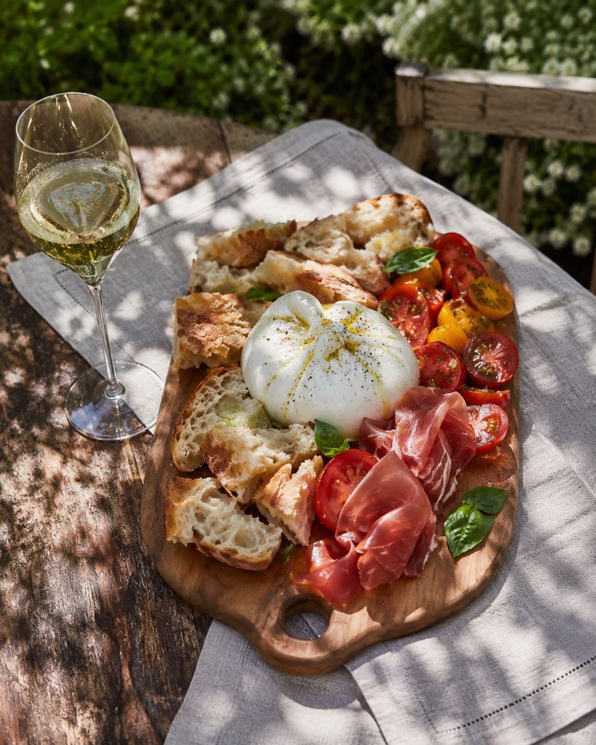 A wooden board with burrata cheese, sliced tomatoes, prosciutto, basil, and torn bread sits on a rustic table next to a glass of white wine, bathed in dappled sunlight.