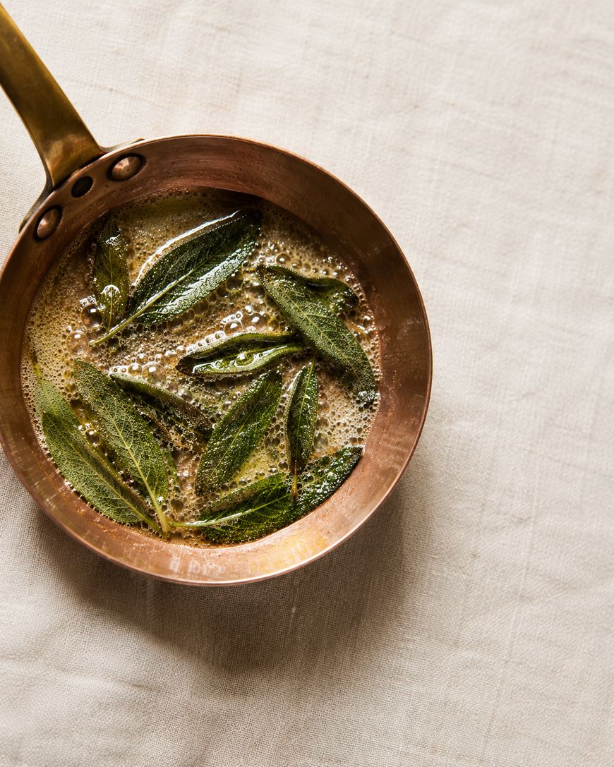 A copper pan with bubbling brown butter and fresh sage leaves sits on a light beige cloth.
