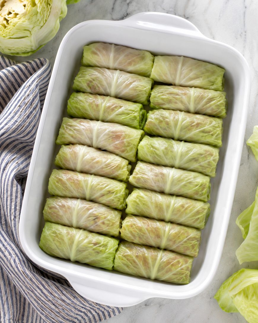 A white baking dish filled with neatly arranged stuffed cabbage rolls, placed on a marble surface next to a striped kitchen towel and loose cabbage leaves.