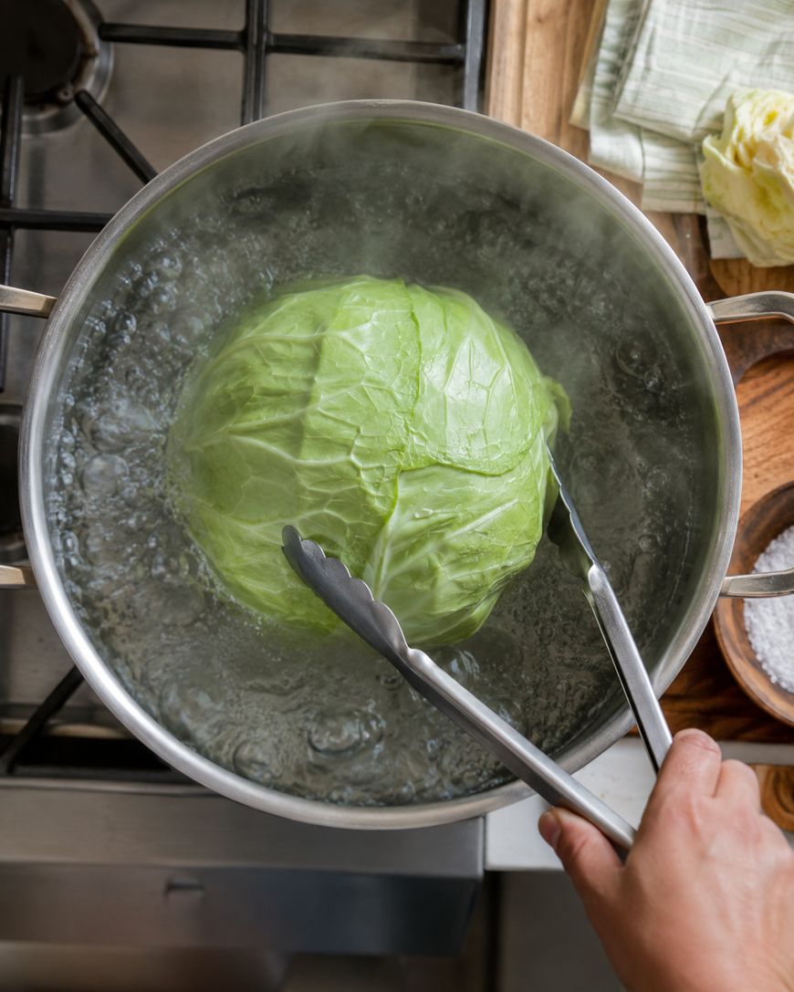 A hand uses metal tongs to hold a whole green cabbage submerged in a pot of boiling water on a stovetop, with steam rising and kitchen items in the background.