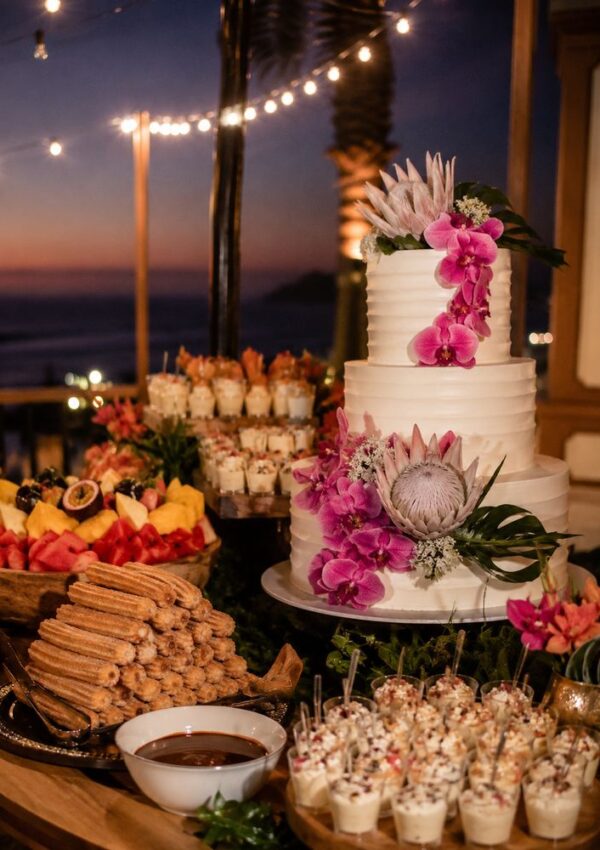 A three-tiered white cake decorated with pink and purple flowers is surrounded by assorted desserts, perfect for a dreamy Cabo wedding or destination celebration, on an outdoor table at sunset adorned with string lights.