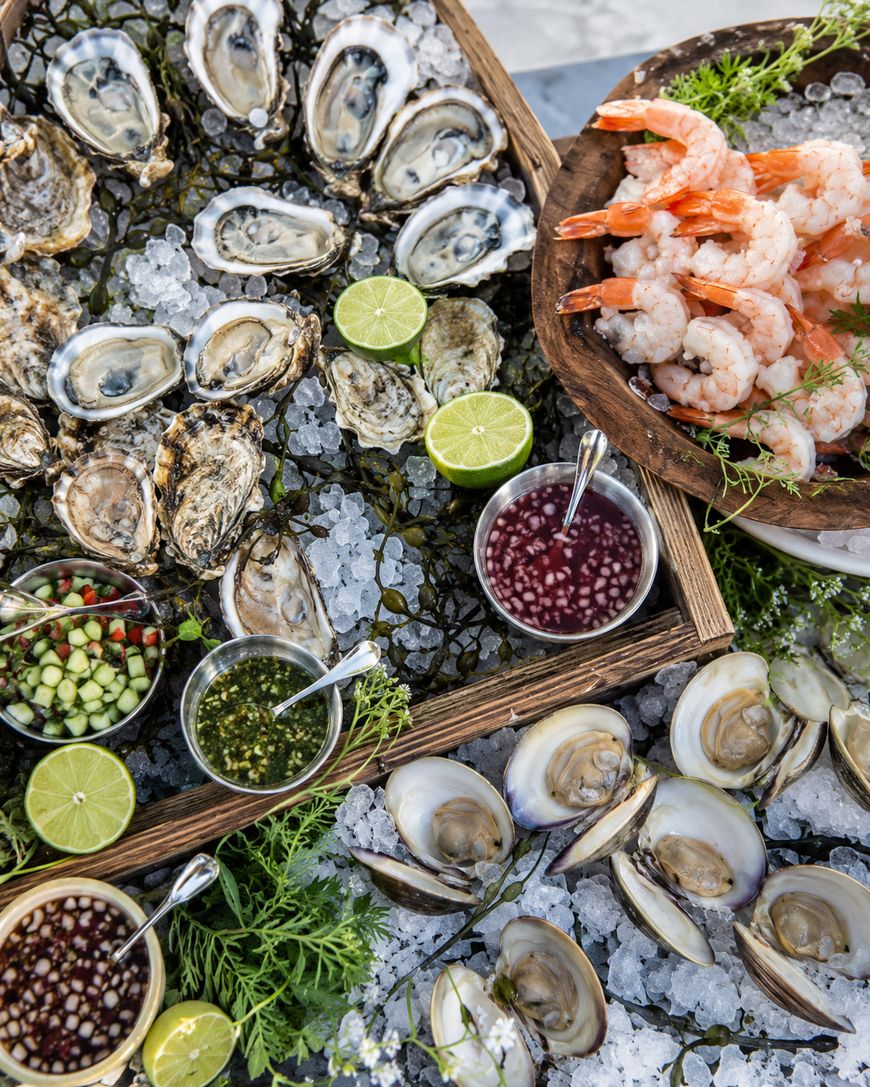 A seafood platter on ice featuring oysters, clams, shrimp, lime halves, and assorted dipping sauces in small bowls, garnished with fresh herbs.