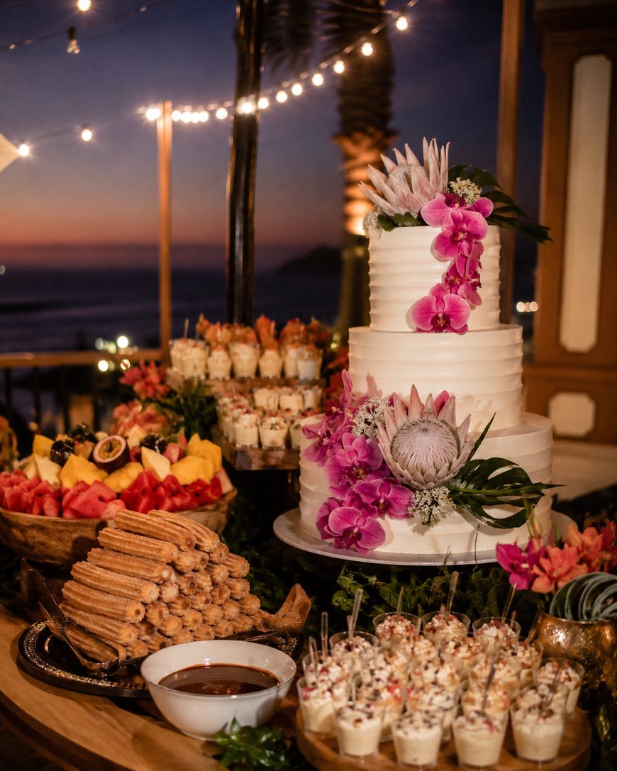 A tiered white cake with pink flowers is surrounded by assorted desserts like churros, fruit, and parfaits on a decorated table outdoors at sunset with string lights and palm trees in the background.
