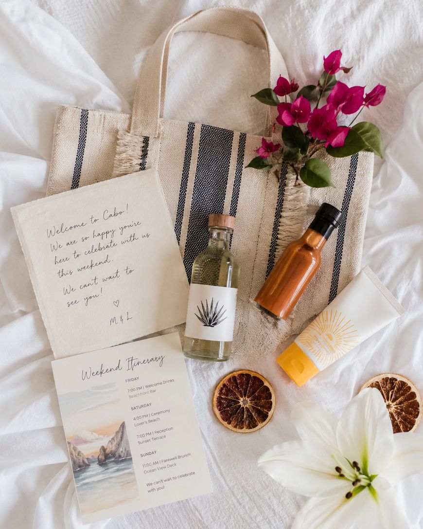 A flat lay of a striped tote bag, two bottles, a tube, pink flowers, dried orange slices, a white lily, a handwritten welcome note, and a printed weekend itinerary on a white cloth background.