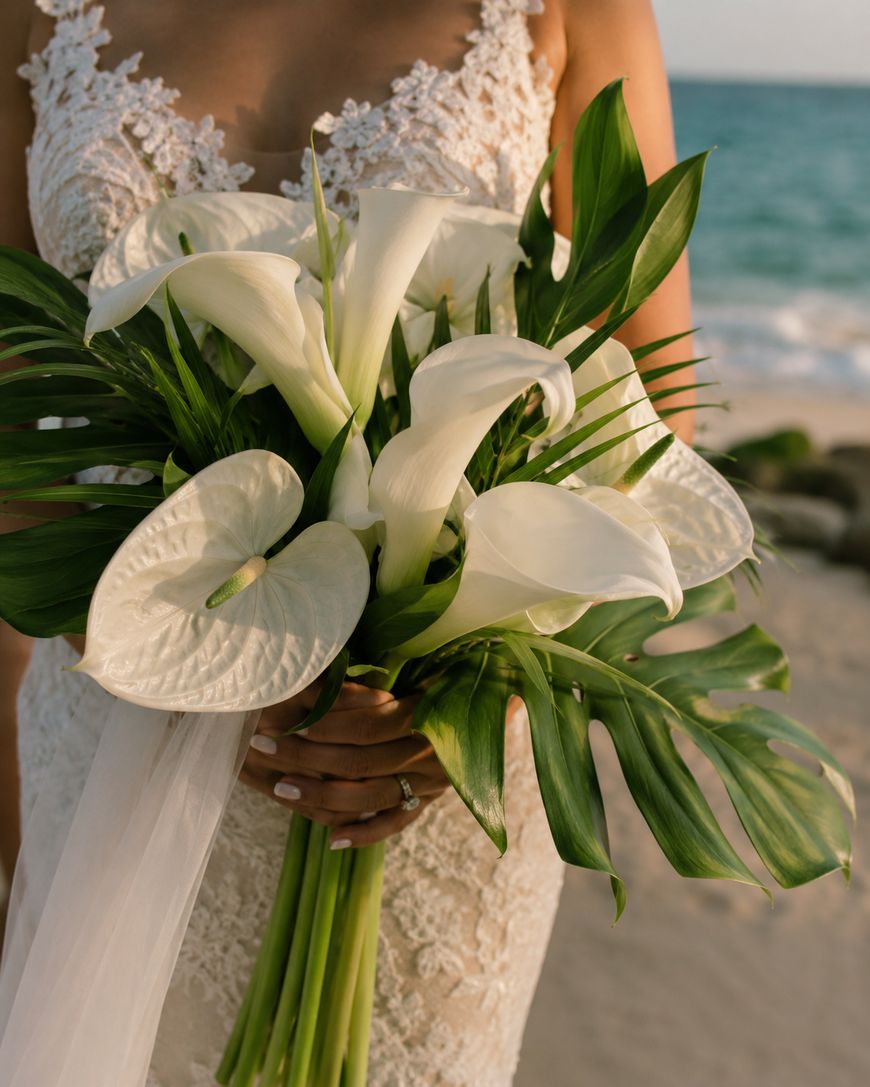 A bride in a lace dress holds a bouquet of white calla lilies, anthuriums, and green tropical leaves on a sandy beach near the ocean.