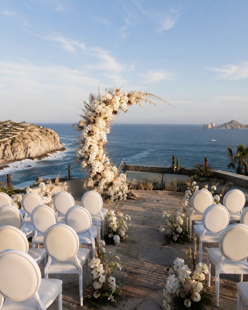 An outdoor wedding ceremony setup overlooks the ocean and cliffs, featuring rows of white chairs and a crescent-shaped floral arch adorned with white flowers under a clear blue sky.