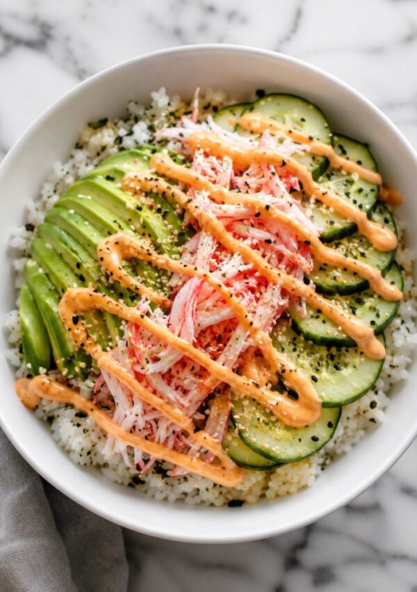 A white bowl filled with sushi rice, sliced avocado, cucumber, imitation crab, black sesame seeds, and a drizzle of spicy orange mayo sauce on top—this california roll bowl is set on a marble surface.