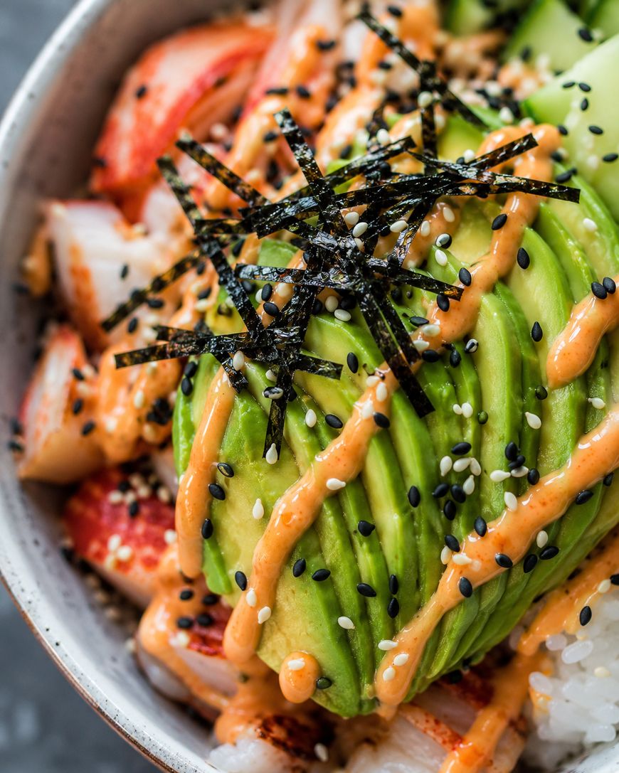 A close-up of a sushi bowl with sliced avocado, crab sticks, cucumber, sushi rice, drizzled spicy mayo, black and white sesame seeds, and shredded nori strips on top.