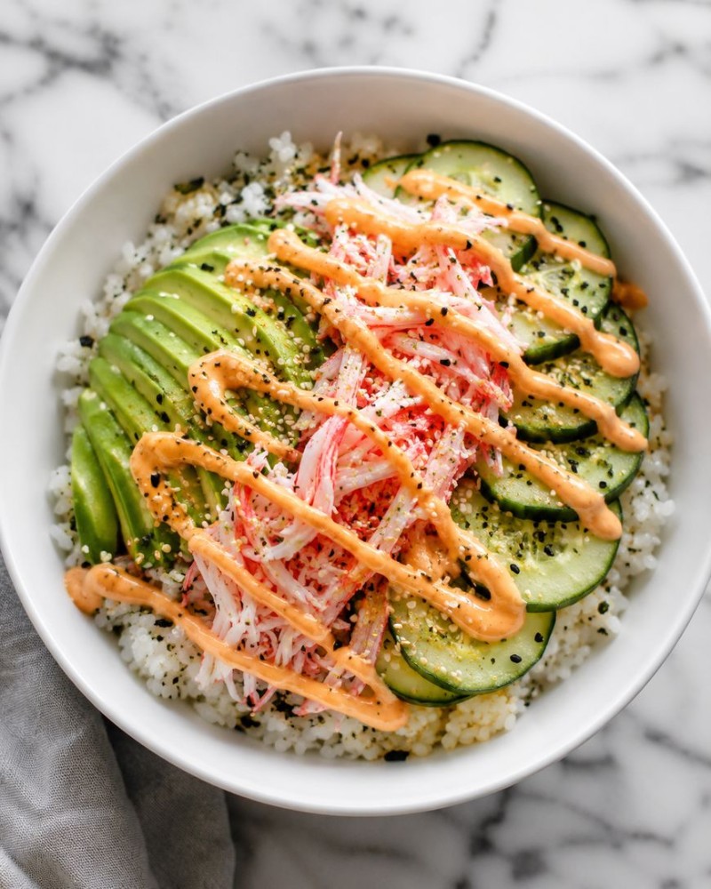 A white bowl filled with sushi rice, sliced avocado, cucumber, imitation crab, black sesame seeds, and a drizzle of spicy orange mayo sauce on top—this california roll bowl is set on a marble surface.