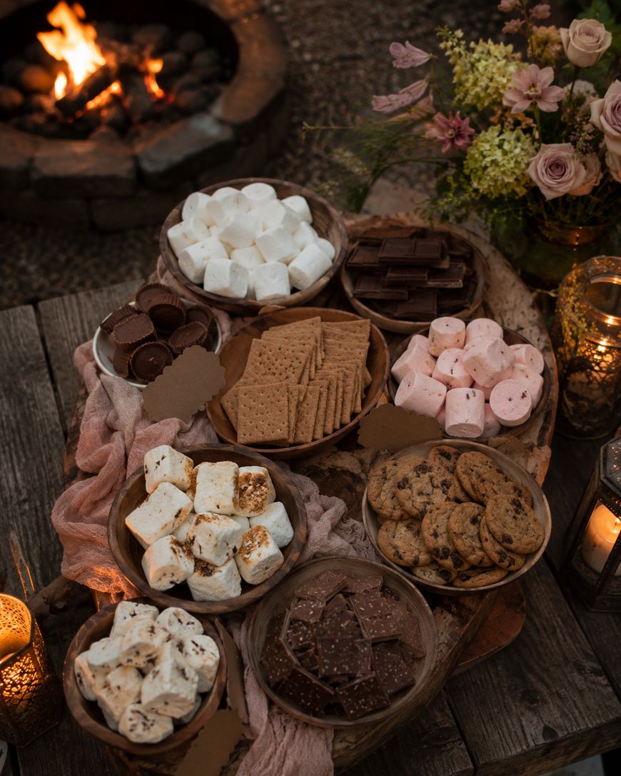 A rustic outdoor table displays bowls of marshmallows, chocolate bars, graham crackers, cookies, and peanut butter cups for s’mores, with flowers, candles, and a firepit glowing in the background.