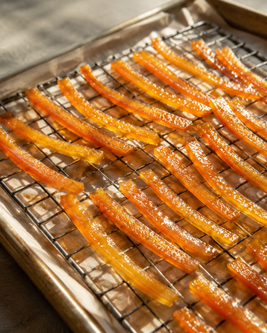 Orange candied citrus peels are arranged in rows on a wire rack set over a parchment-lined baking sheet, with sunlight illuminating the glossy surface of the peels.