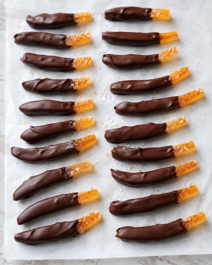 Candied orange peels partially dipped in dark chocolate, arranged in neat rows on a sheet of parchment paper. The sugared ends are exposed, contrasting with the glossy chocolate coating.