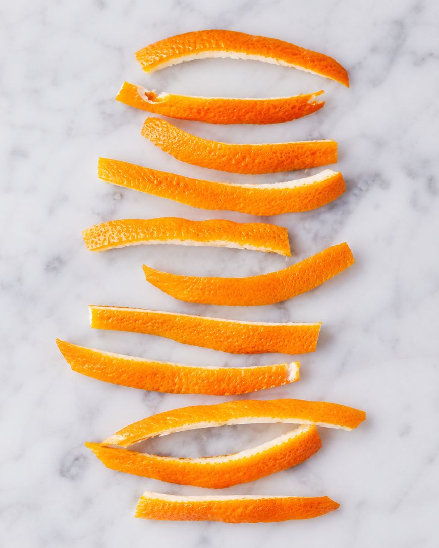 Ten orange peel strips are neatly arranged in parallel rows on a white marble surface. The strips display the vibrant orange exterior and some white pith underneath.
