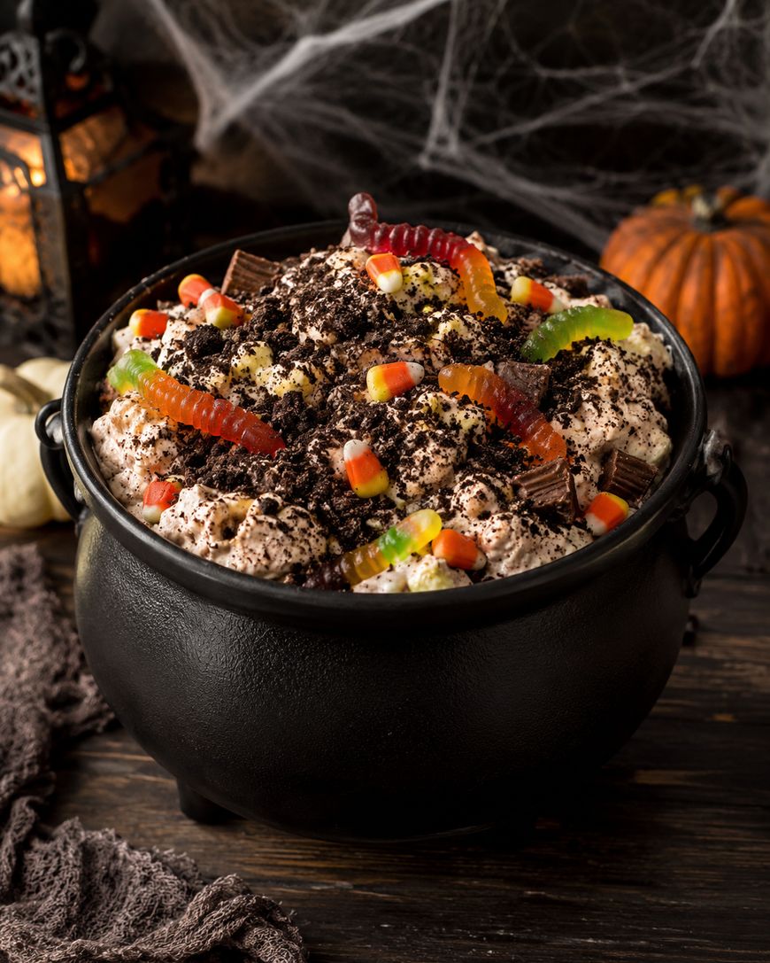 A black cauldron filled with Halloween-themed dessert topped with crumbled cookies, candy corn, and gummy worms. In the background are cobweb decorations, a pumpkin, and a lantern.