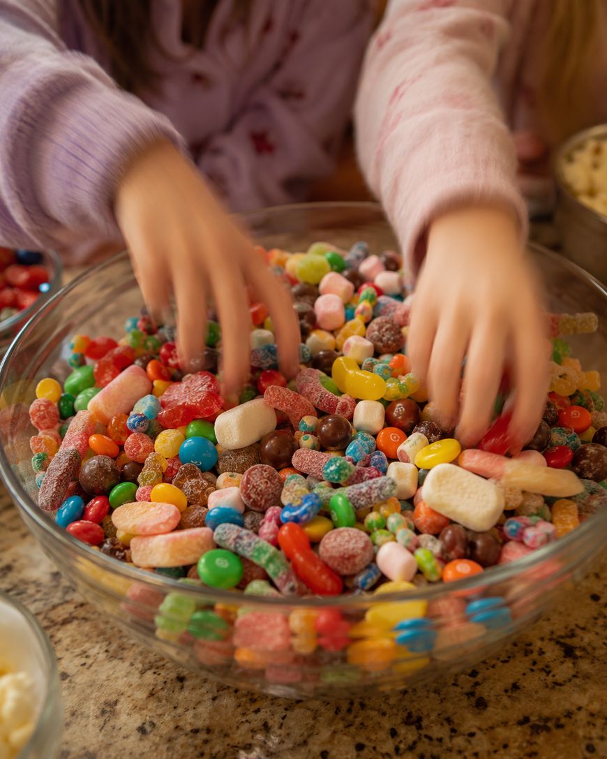 Two children reach into a large glass bowl filled with assorted colorful candies and gummies, set on a kitchen counter. Only their hands and part of their arms are visible.