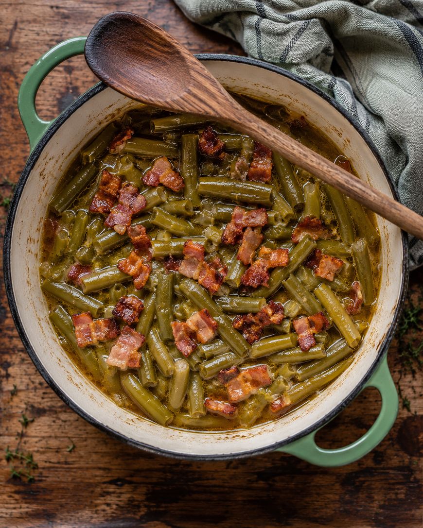 A pot of cooked green beans topped with crispy bacon pieces, sitting on a wooden table with a wooden spoon resting on the pot’s edge and a striped cloth nearby.